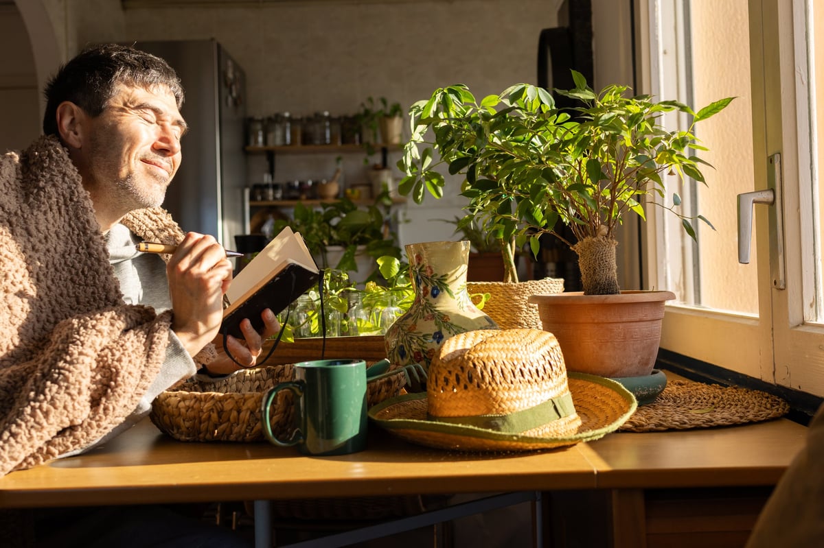 Mature man wrapped in a cozy blanket writing in a journal at a wooden table, bathed in morning sunlight, peaceful self-care moment