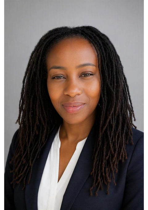 Professional headshot of a woman with long dark locs, wearing a black blazer and white shirt against a gray background