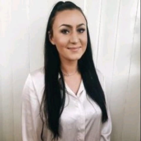 Woman with long black hair wearing a white button-up shirt, smiling at the camera in an indoor setting